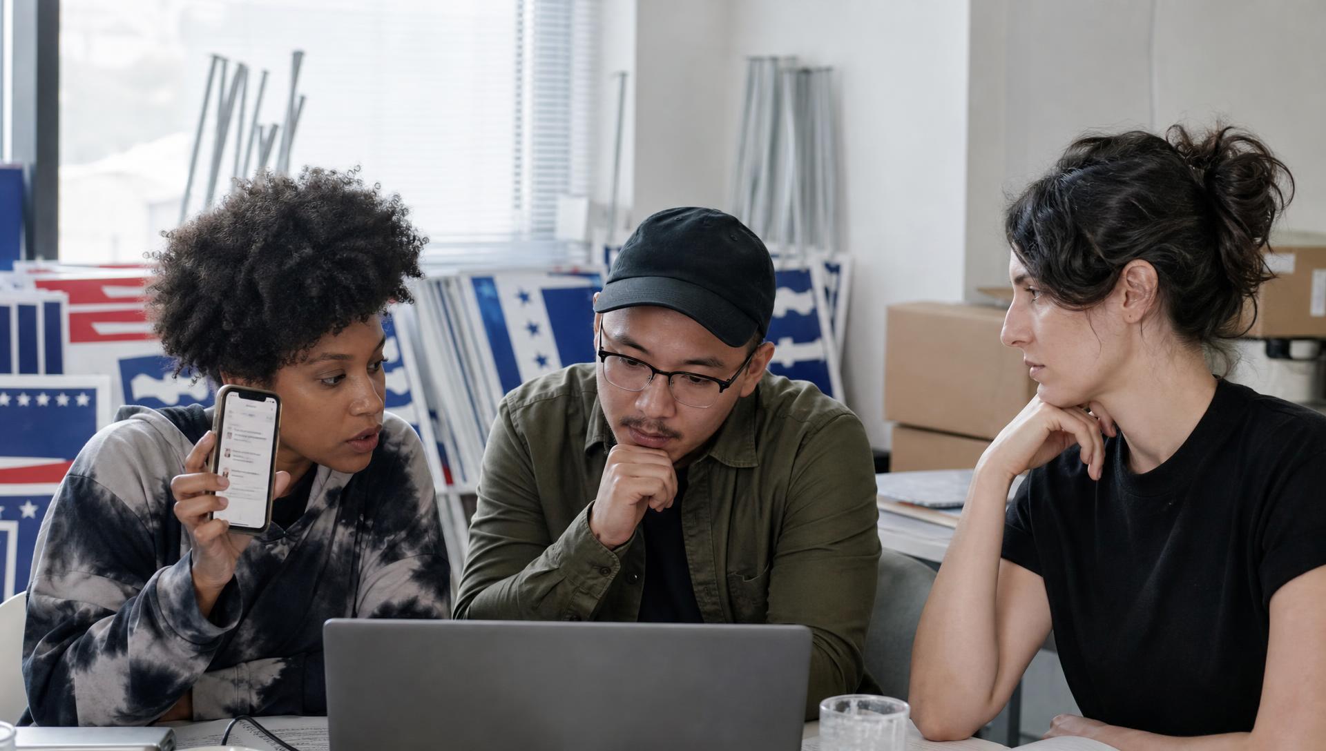 Three campaign researchers reviewing a candidate website on a phone and laptop in an office stacked with yard signs